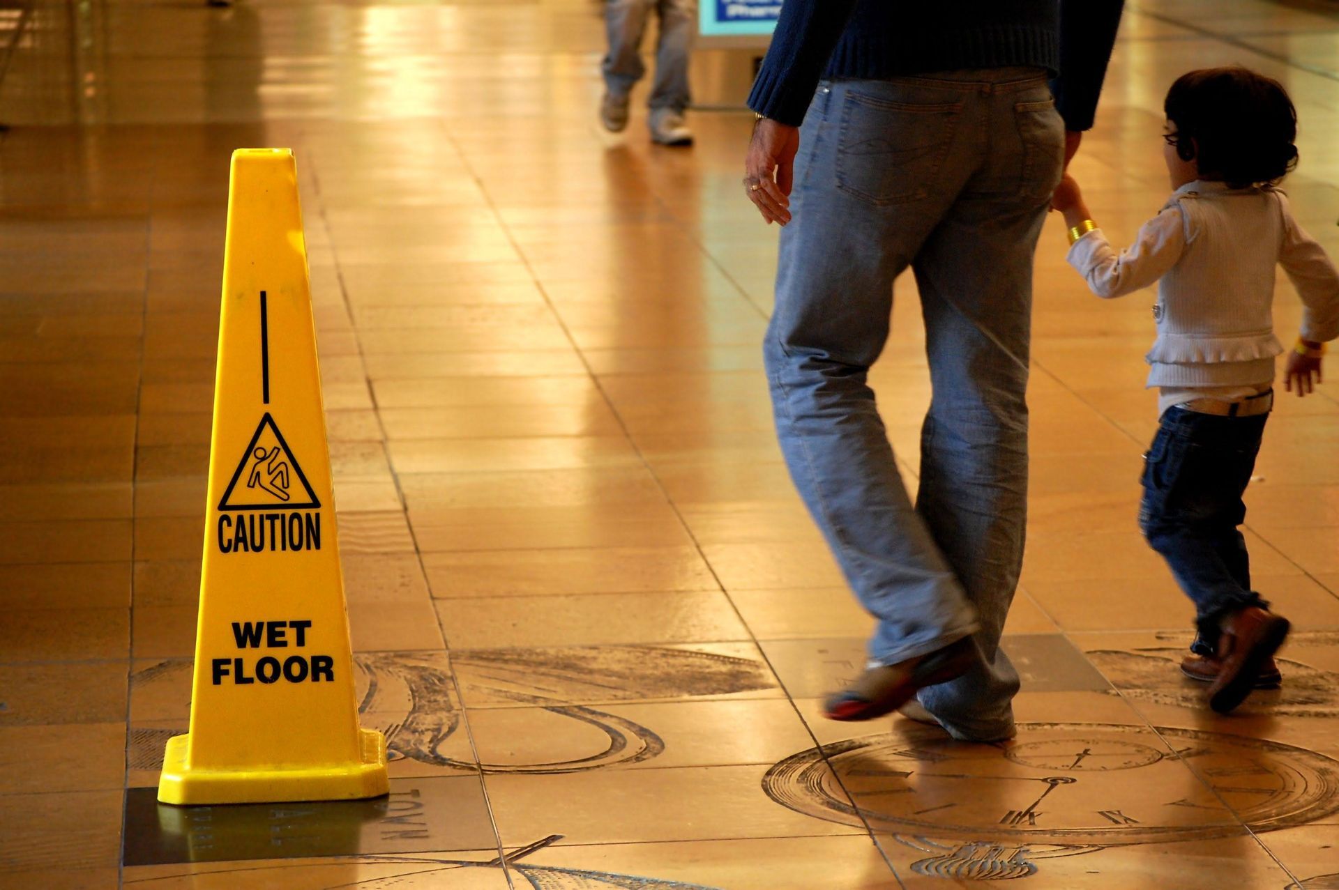 Yellow caution cone warns of wet floor. A person holds a child's hand, walking on a reflective floor.