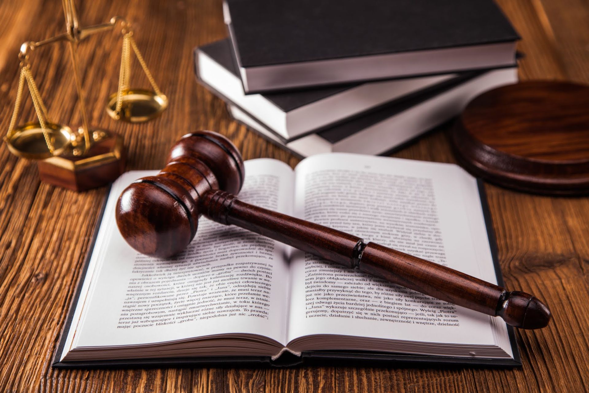Wooden gavel on open book with law books and scales on a wooden table.