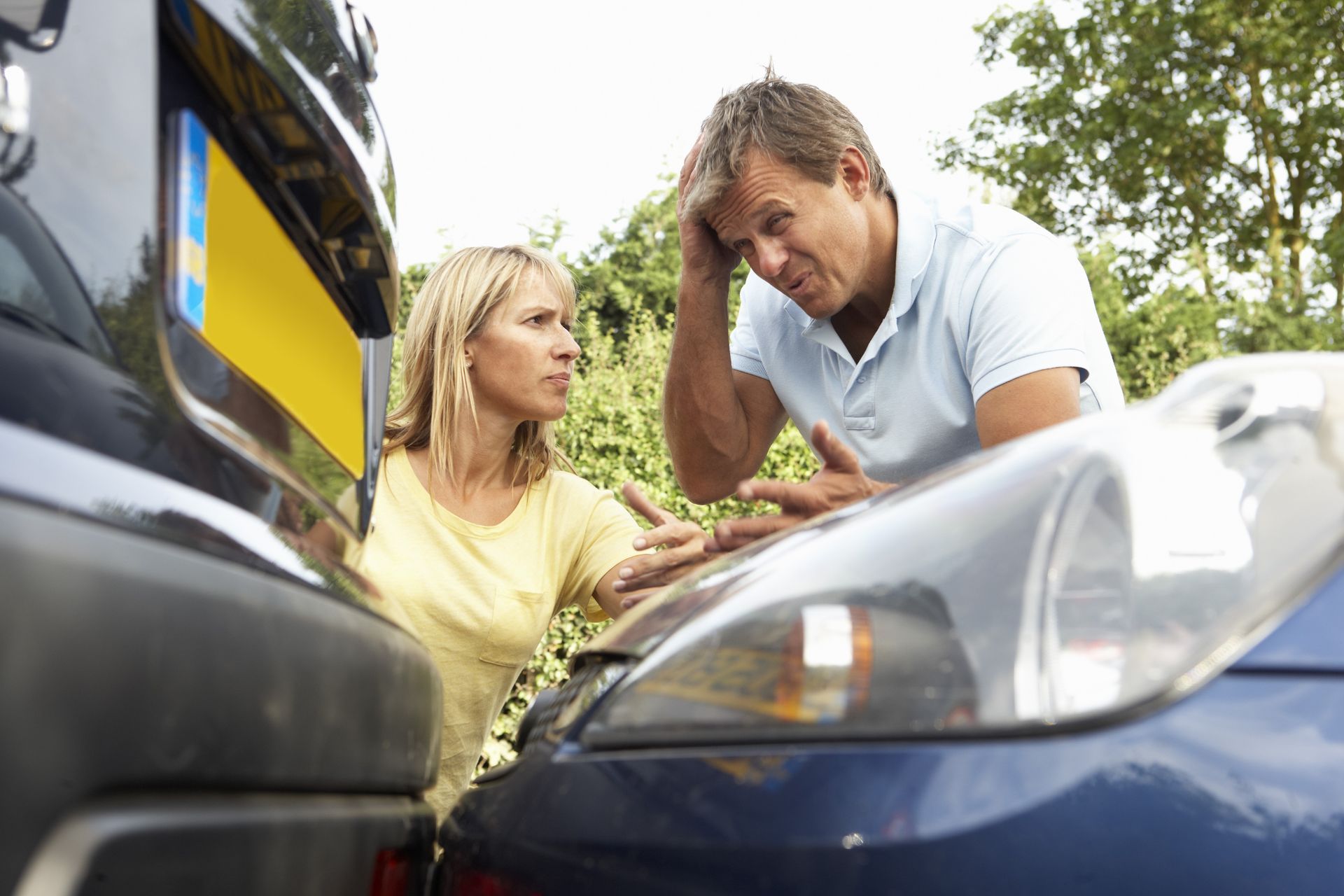 A woman and man assess damage to their cars after a collision; both look concerned, outdoor setting.