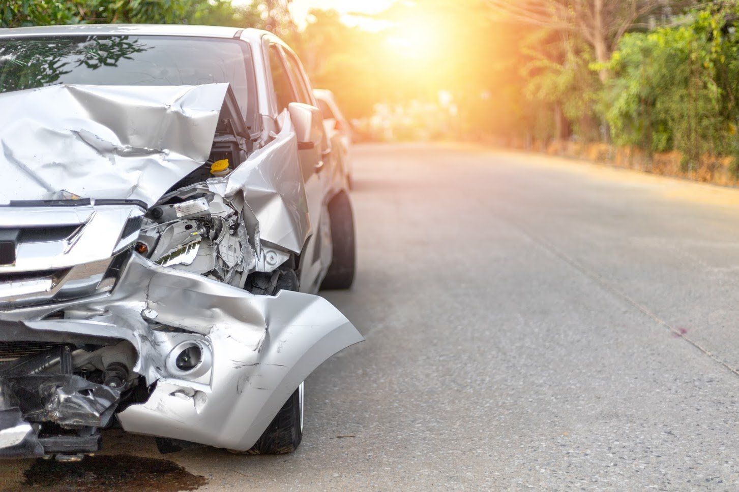 A silver car severely damaged in a crash, on a road with bright sunlight.