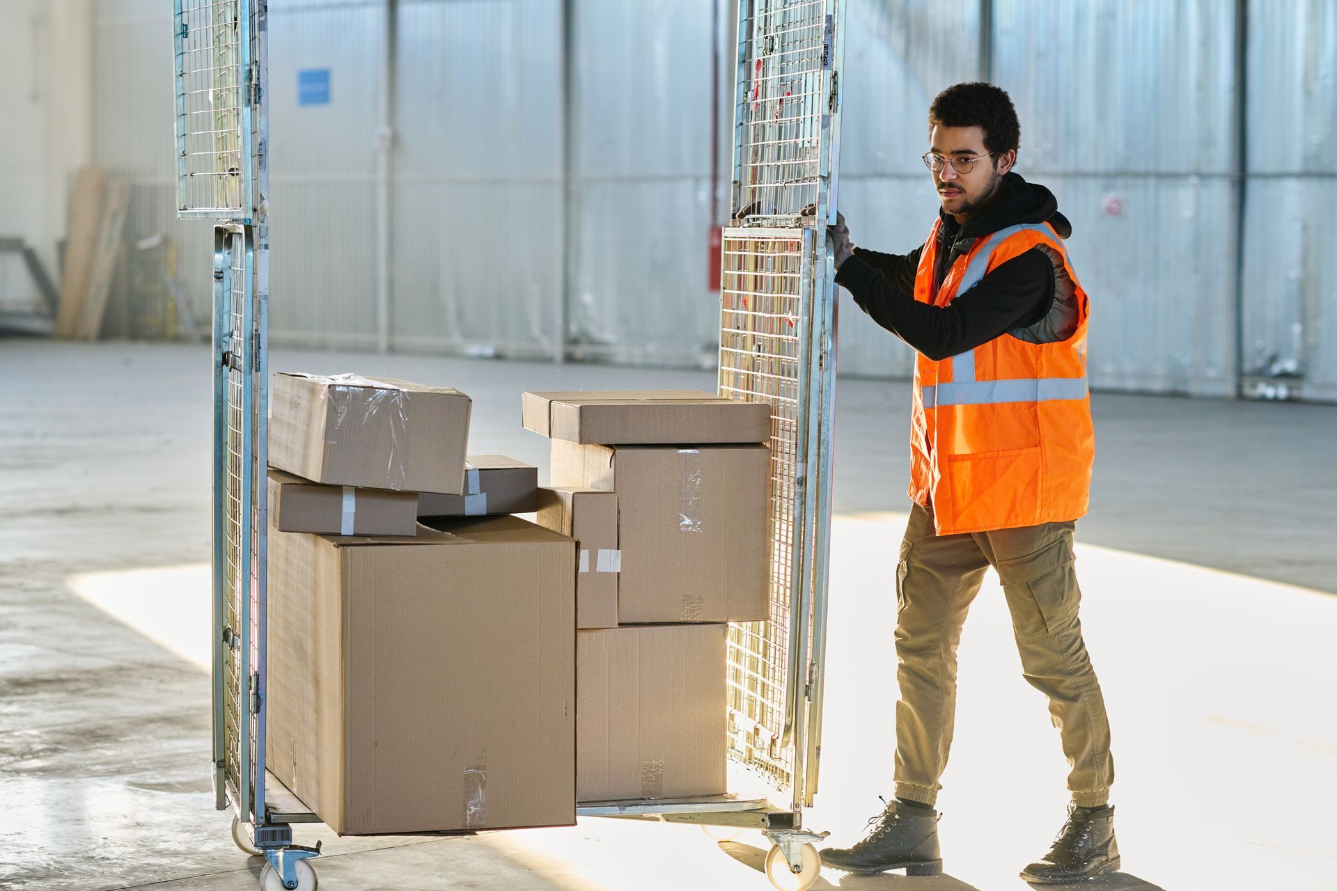 Worker in orange safety vest using a tablet beside stacked cardboard boxes in a warehouse