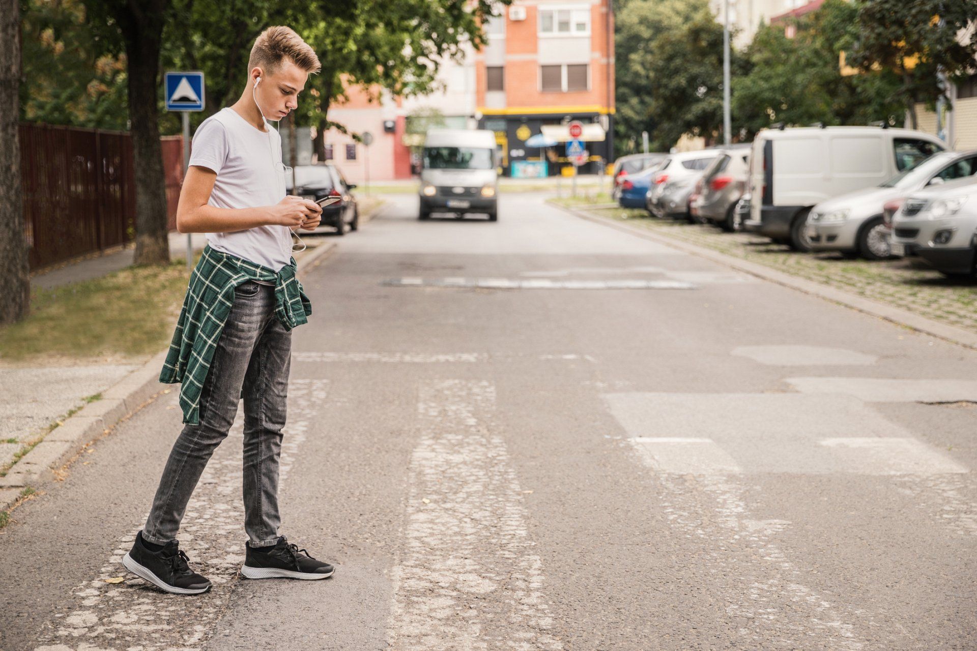 Teenager crossing street, wearing earbuds, looking at phone. Green plaid shirt tied around waist. Cars parked on side.