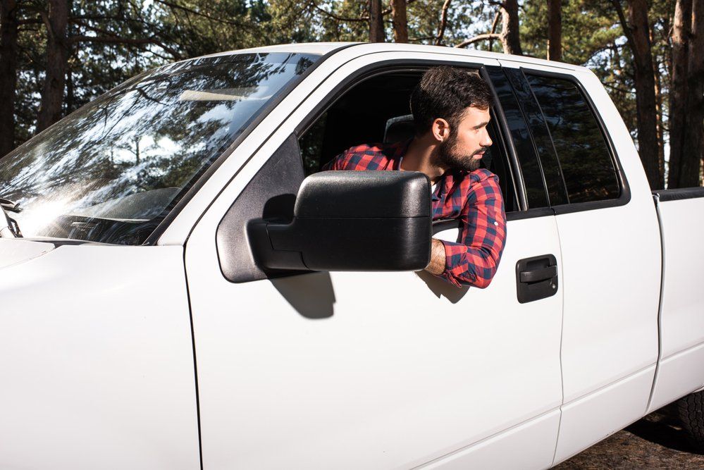 Man in red plaid shirt looking out of the window of a white pickup truck in a forest.
