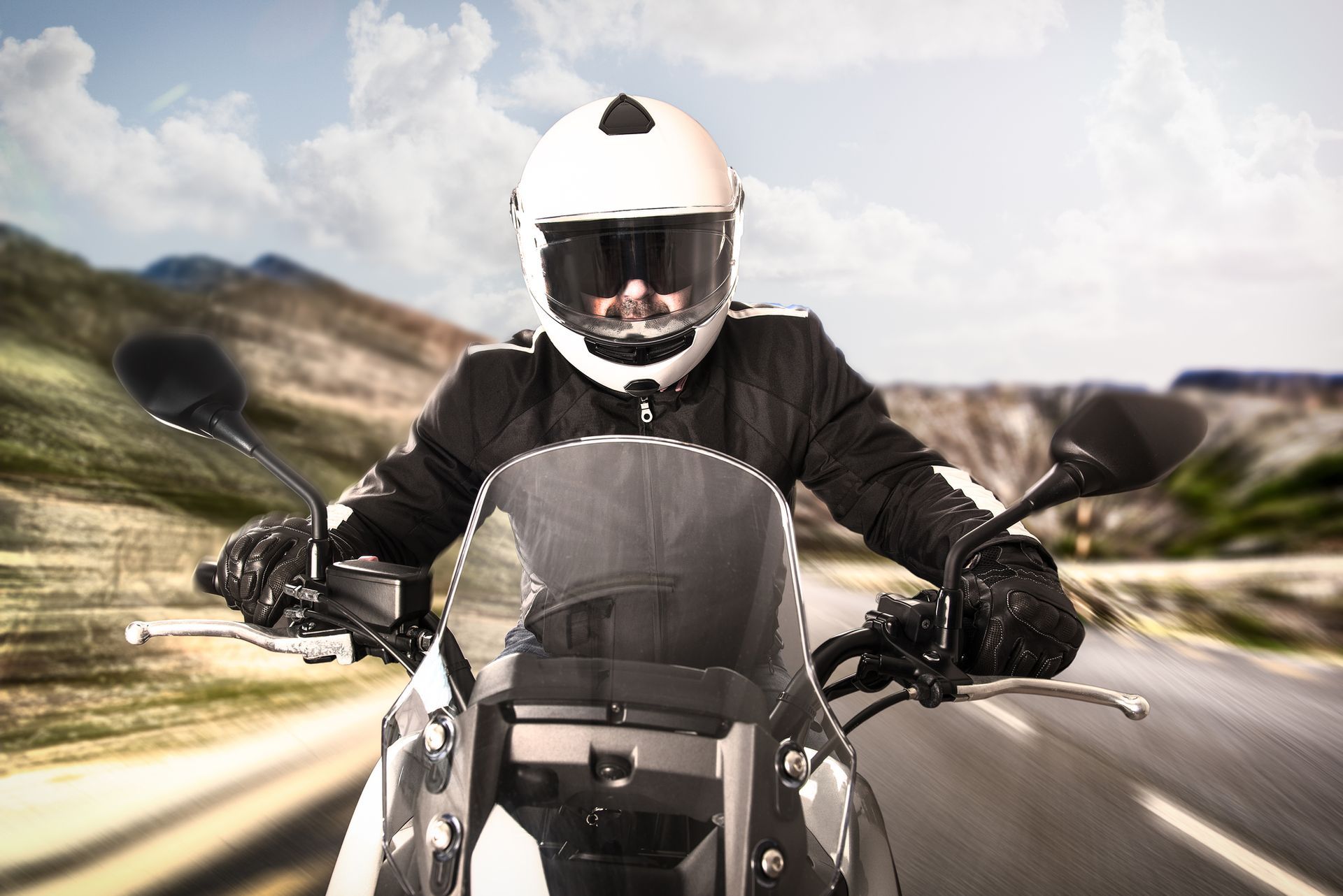 Motorcyclist in white helmet and black jacket riding on a road, mountains in the background.