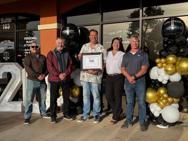 Five people posing outside a storefront, one holding a framed certificate beside black, gold, and white balloons.
