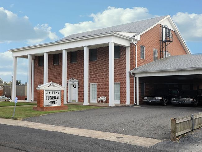 Brick church with white columns and covered entrance, viewed from a gravel lot under a blue sky.