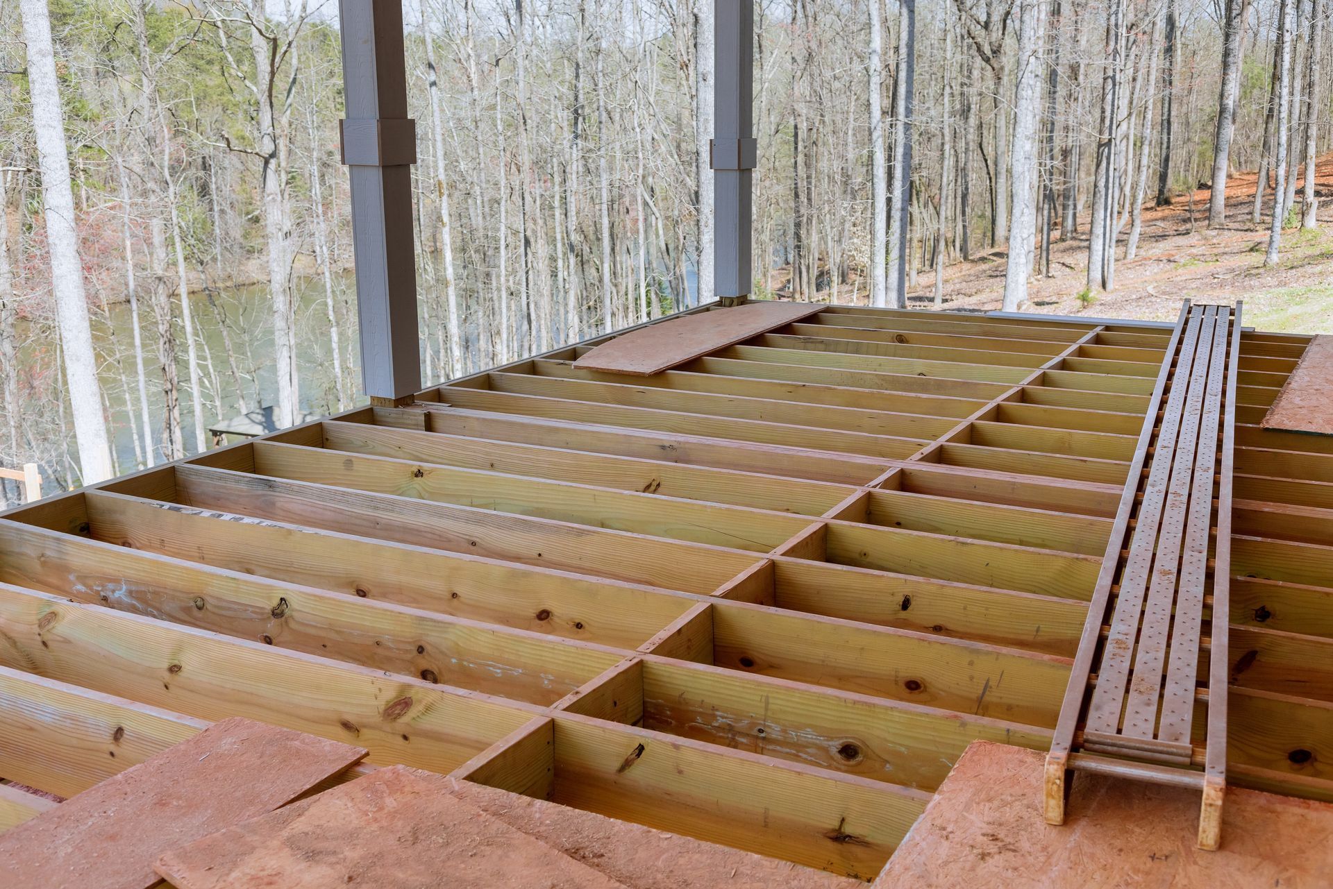 Unfinished wooden deck framing on a screened porch overlooking leafless trees outdoors