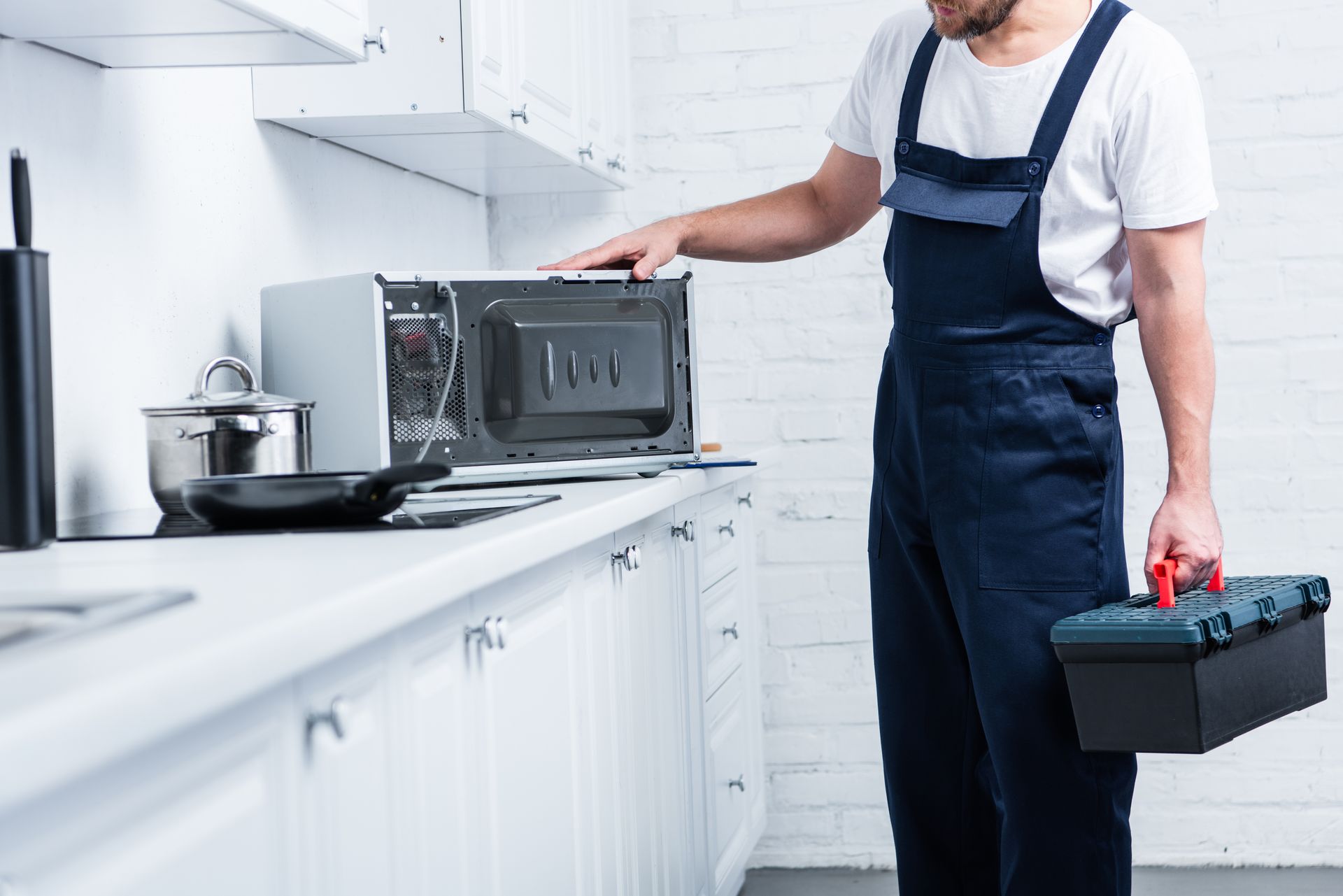 Man in blue overalls with toolbox examining a microwave on a kitchen counter.
