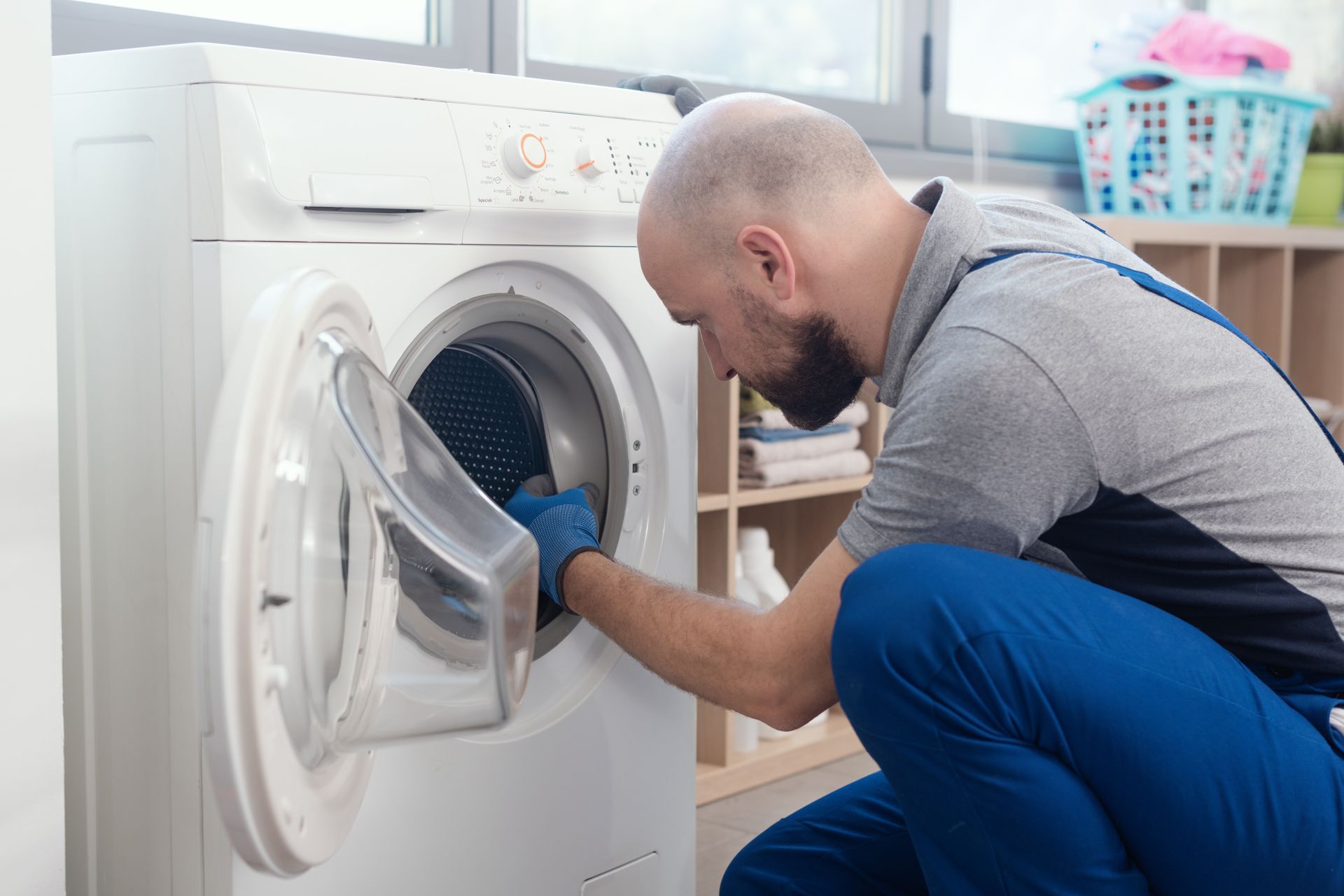 Man in blue overalls inspecting a washing machine, wearing gloves.