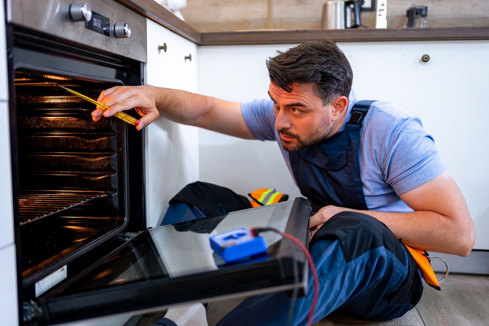 Repairman checks an oven with a tool; he is kneeling, focused, in a kitchen setting.