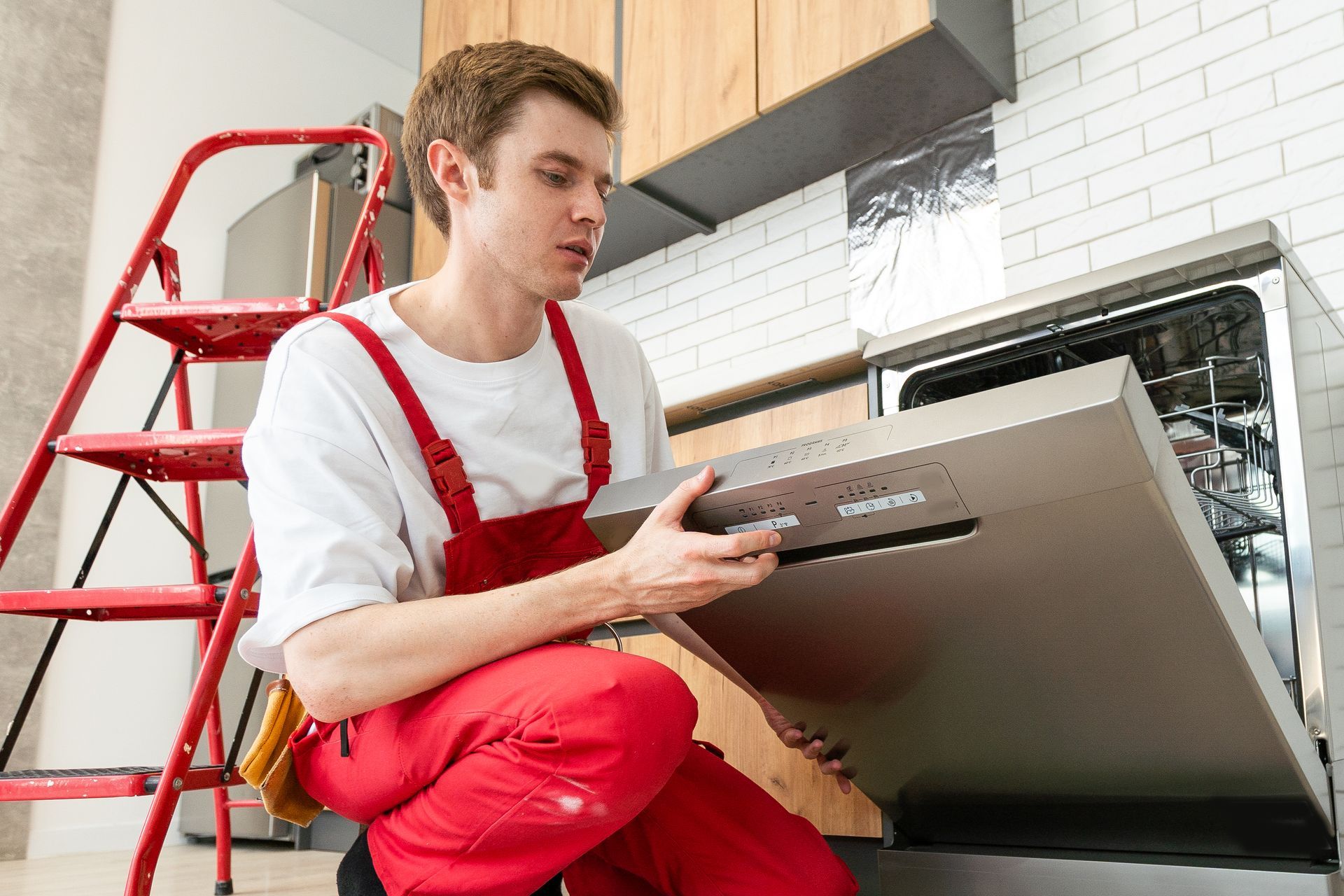 A person in red overalls installs a dishwasher in a kitchen, holding the door panel. A ladder is nearby.
