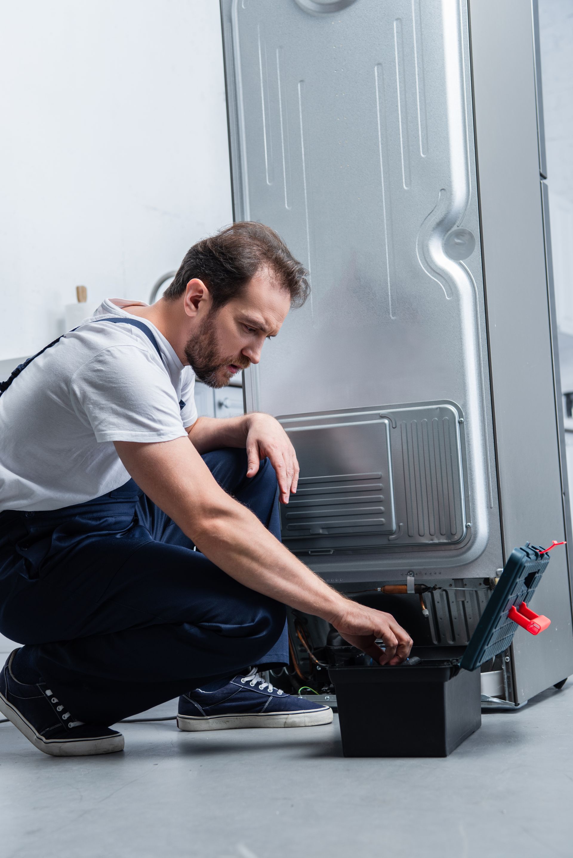 Man in blue overalls kneels, working on the back of a refrigerator, tools in a black box.