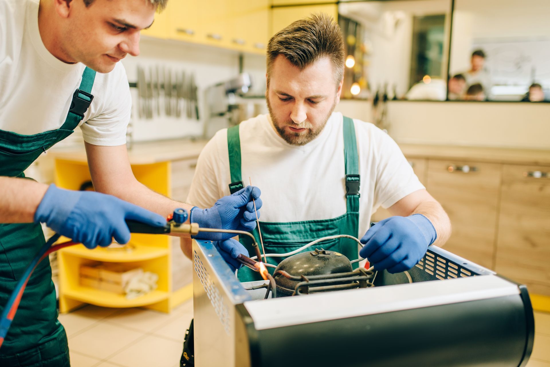 Two technicians in green overalls repairing a refrigerator, one using a blowtorch.