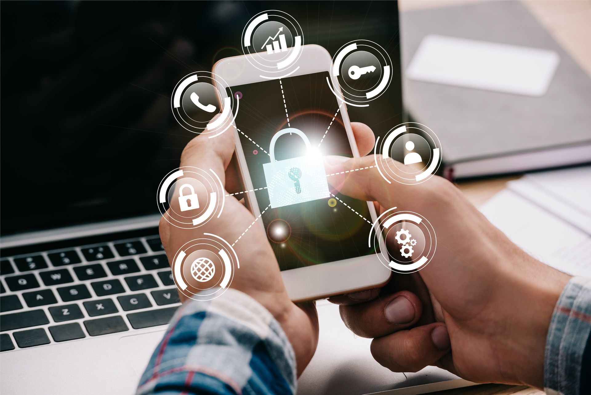Manos sosteniendo un teléfono inteligente con un icono de candado e iconos de seguridad sobre el teclado de una computadora portátil.