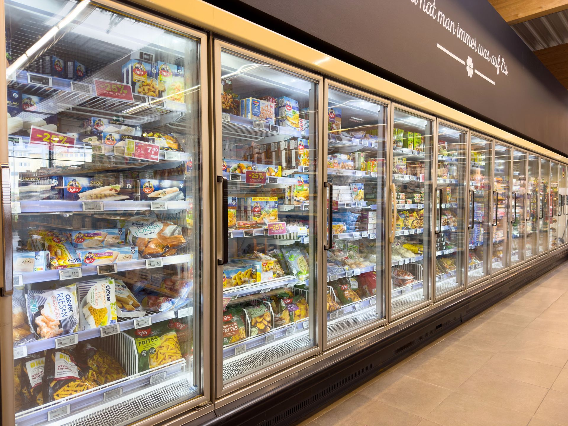 Pasillo refrigerado de comestibles con filas de congeladores con puertas de vidrio repletos de alimentos congelados envasados.