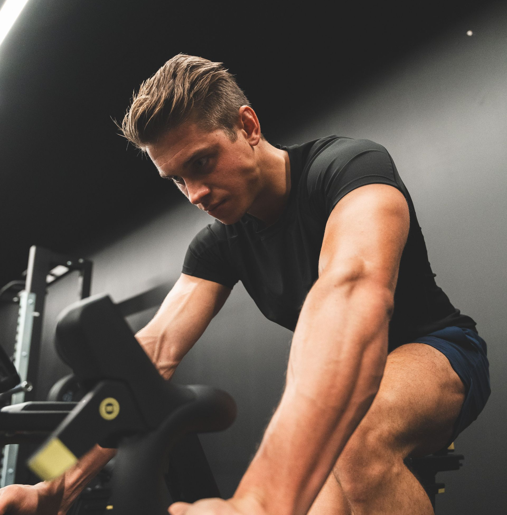 Man exercising on a stationary bike in a dim gym, leaning forward and gripping the handlebars.