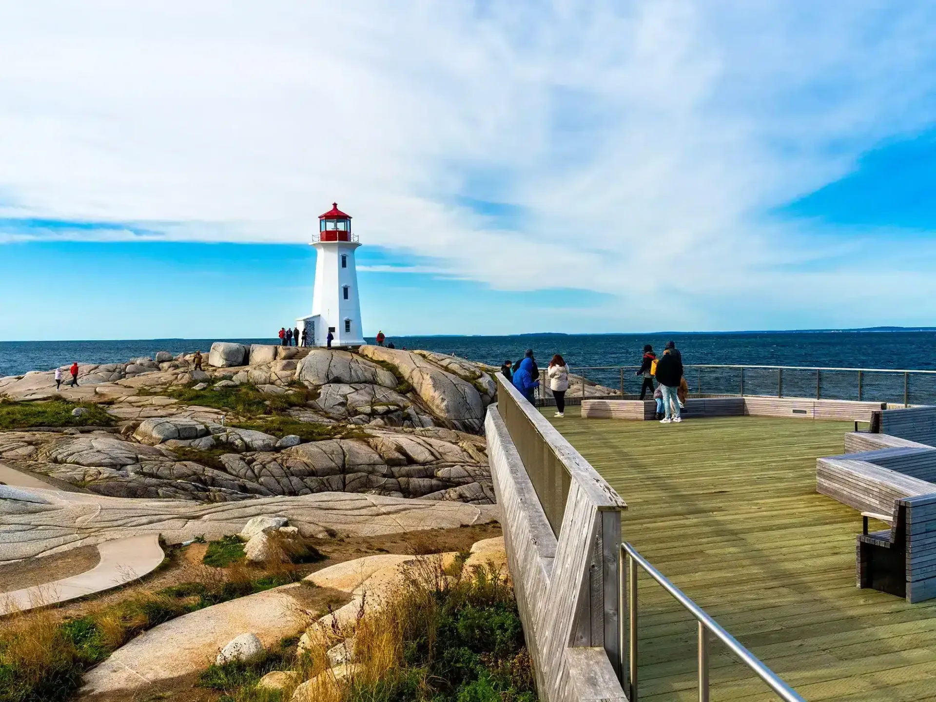 Lighthouse on rocky coast with visitors on a walkway under a bright blue sky