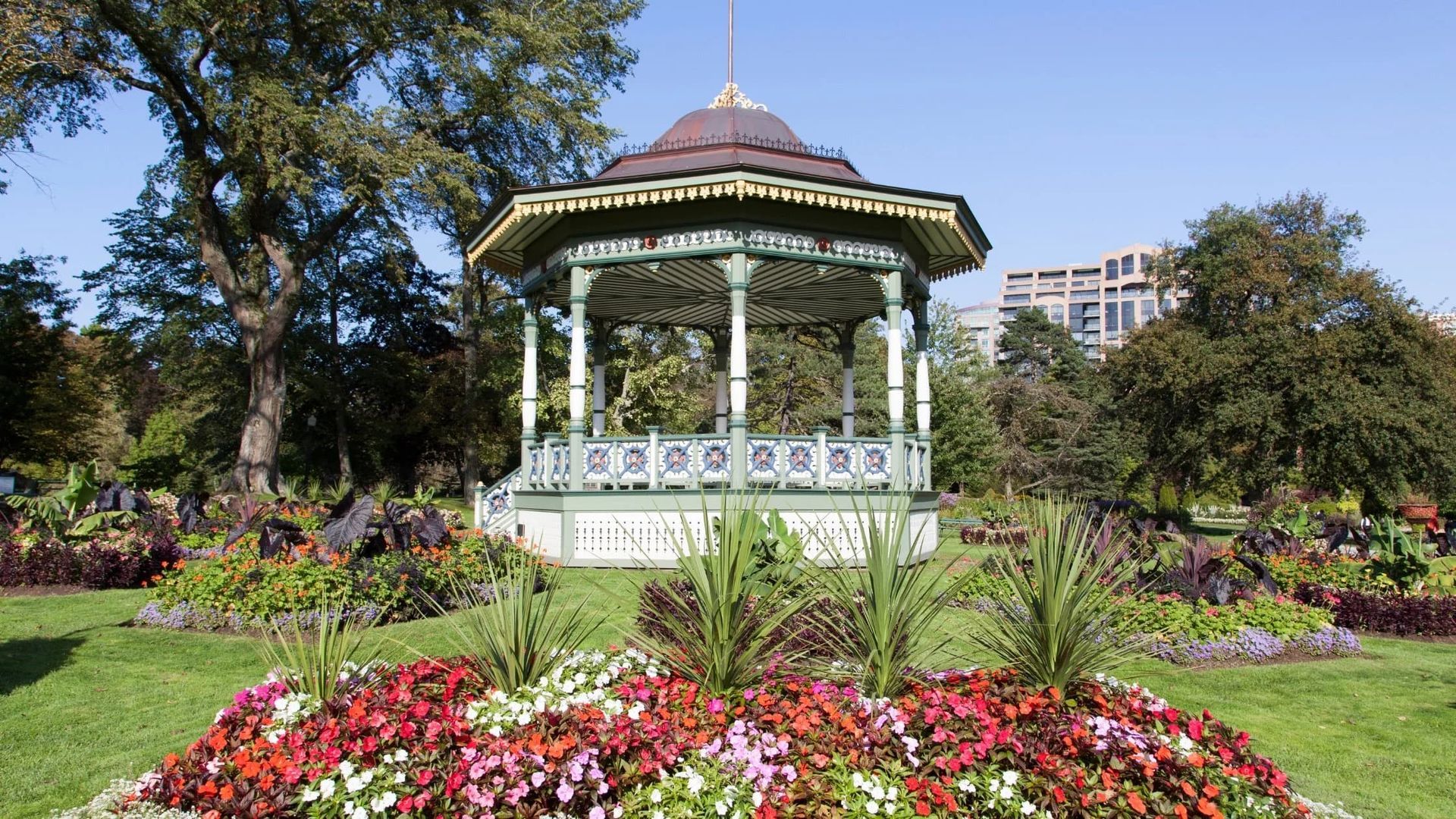 White gazebo in a colorful flower garden with trees and city buildings in the background