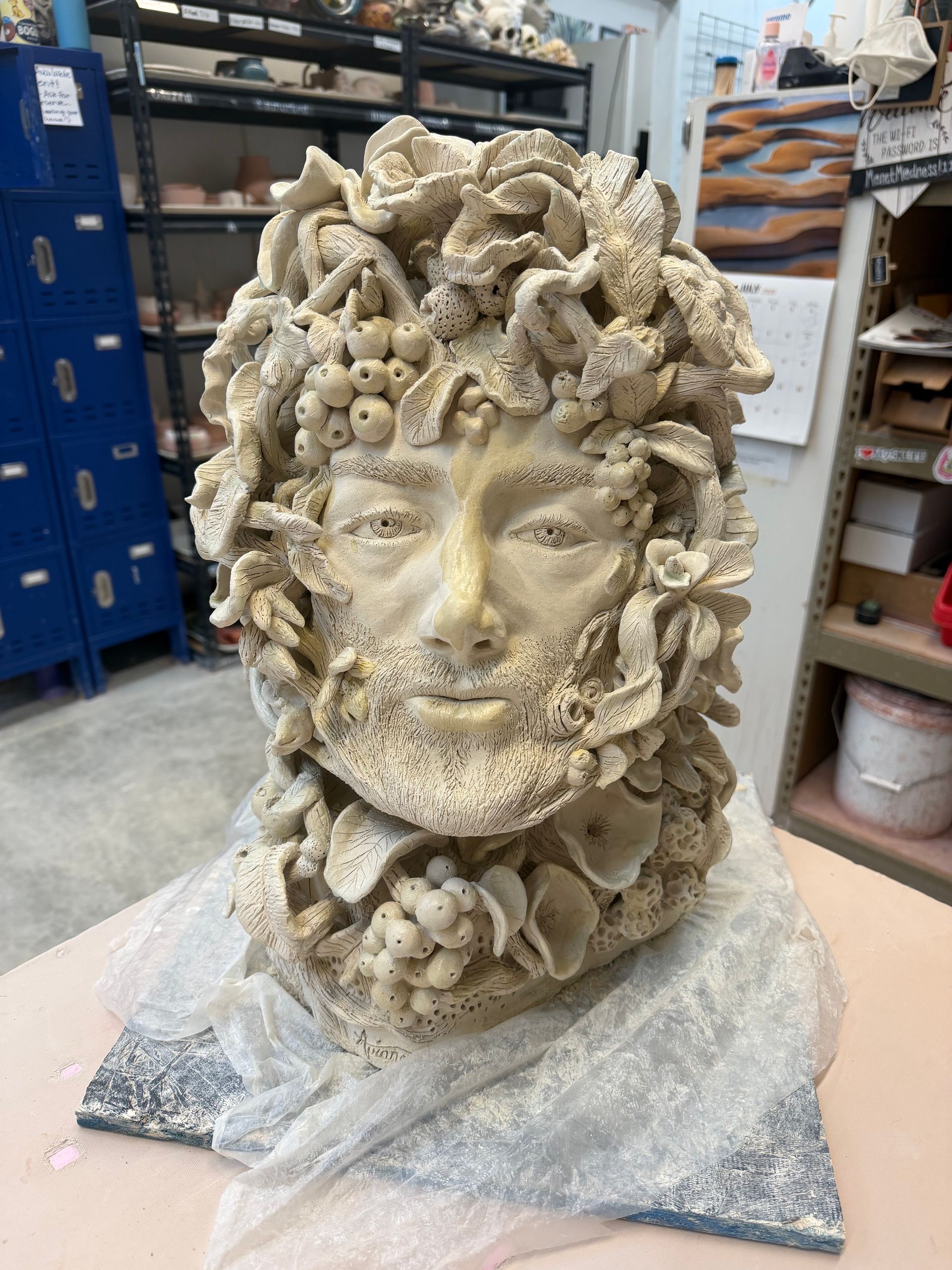 Stone bust with a face made of shells or coral, wrapped in plastic on a workshop table.