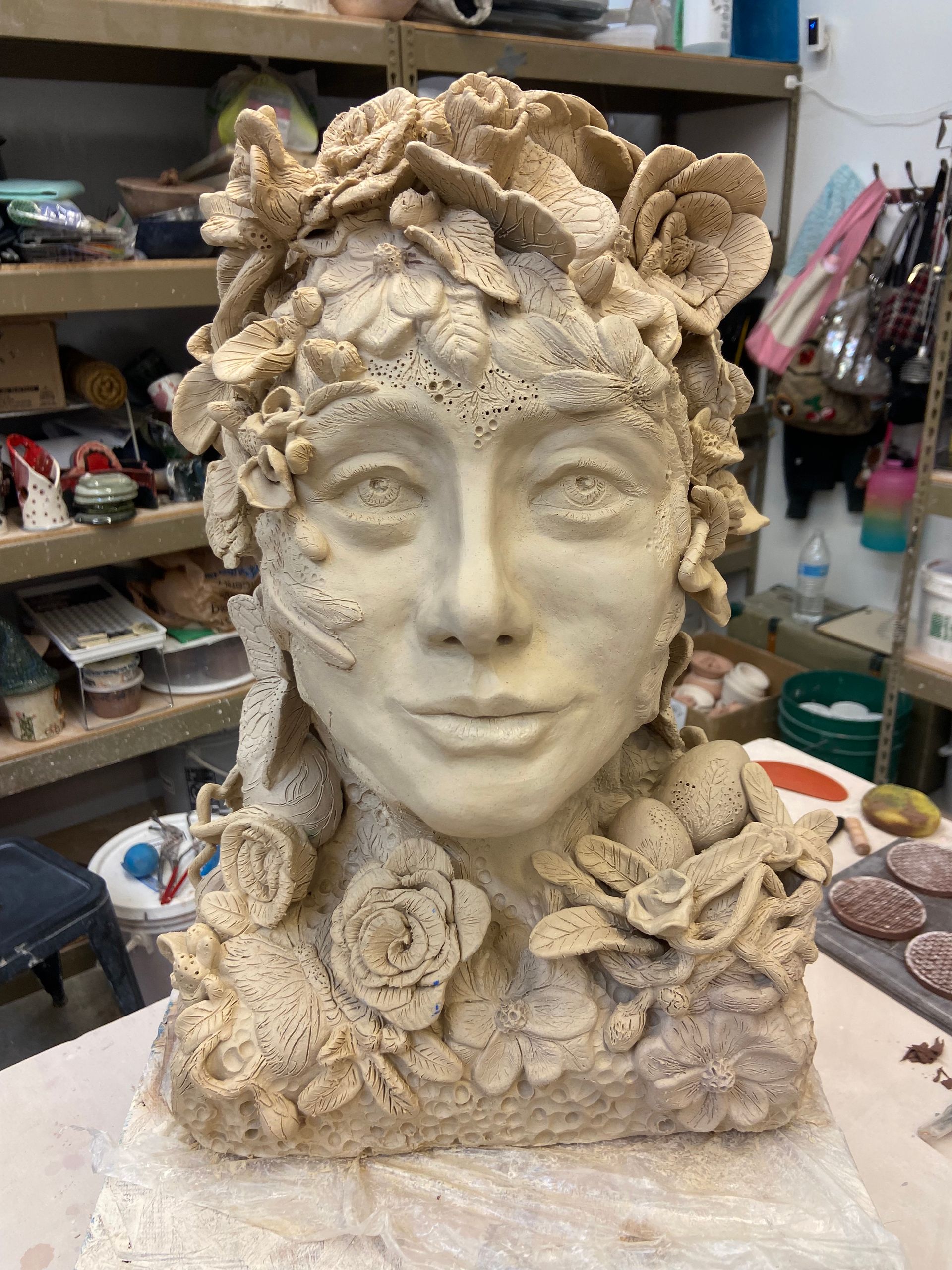 Sculpted ceramic bust with a serene face and floral textures, displayed in a studio workspace.