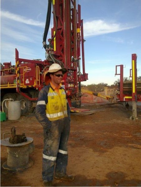 Worker in overalls and hard hat stands near a red drilling rig on a construction site, eyes shielded by sunglasses.