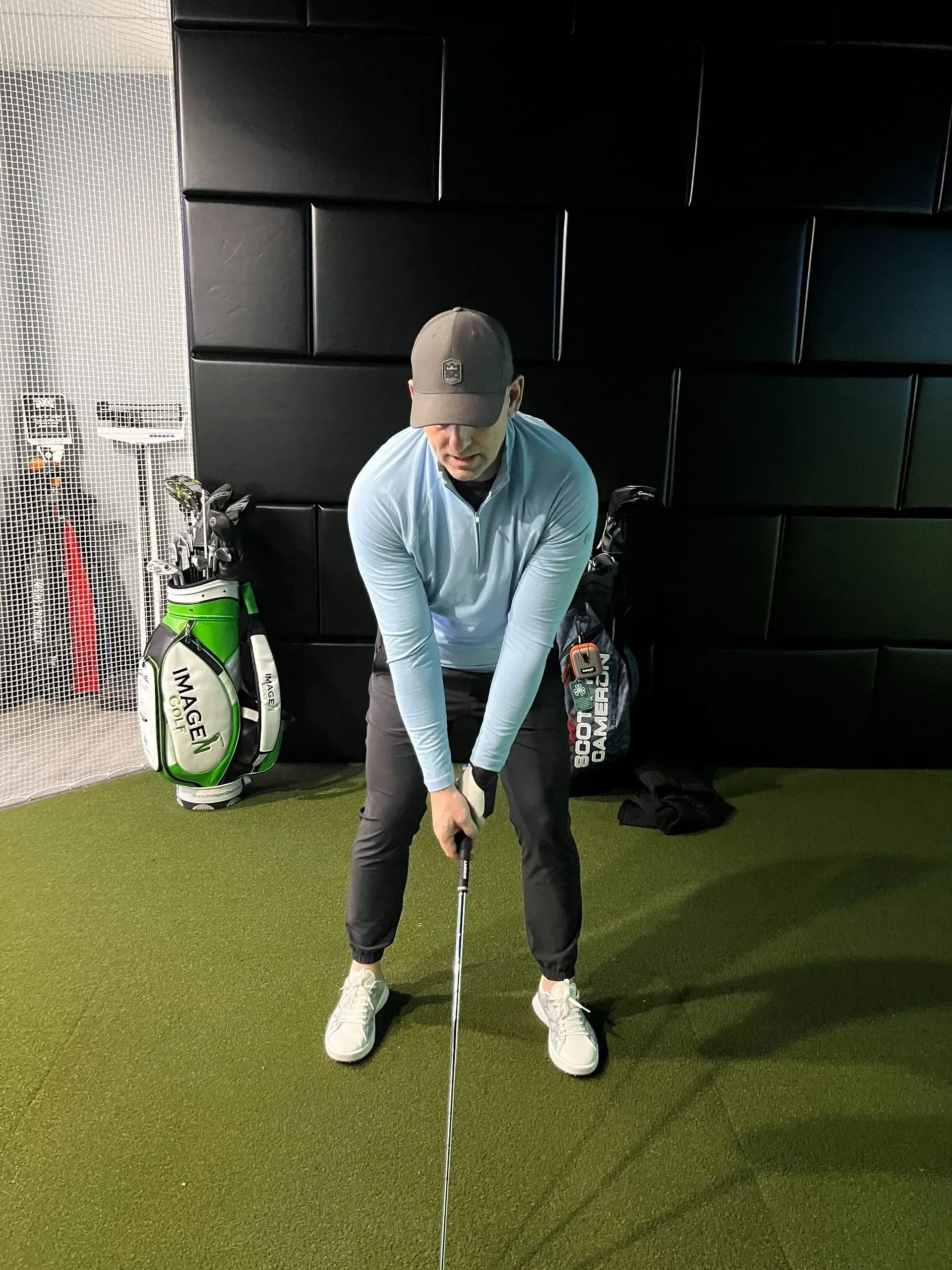 Golfer addressing the ball indoors on green turf, holding a club in a practice stance.