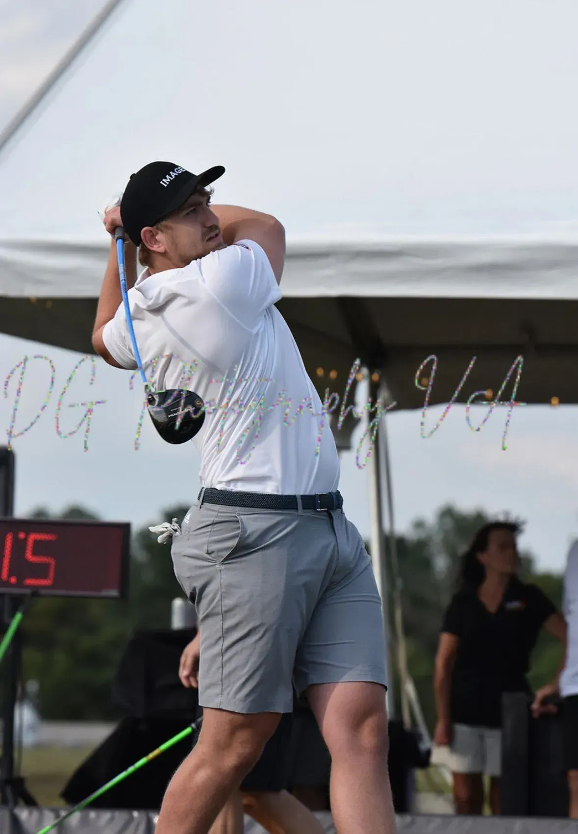 Golfer mid-swing on a course, wearing a white shirt and gray shorts under a canopy.