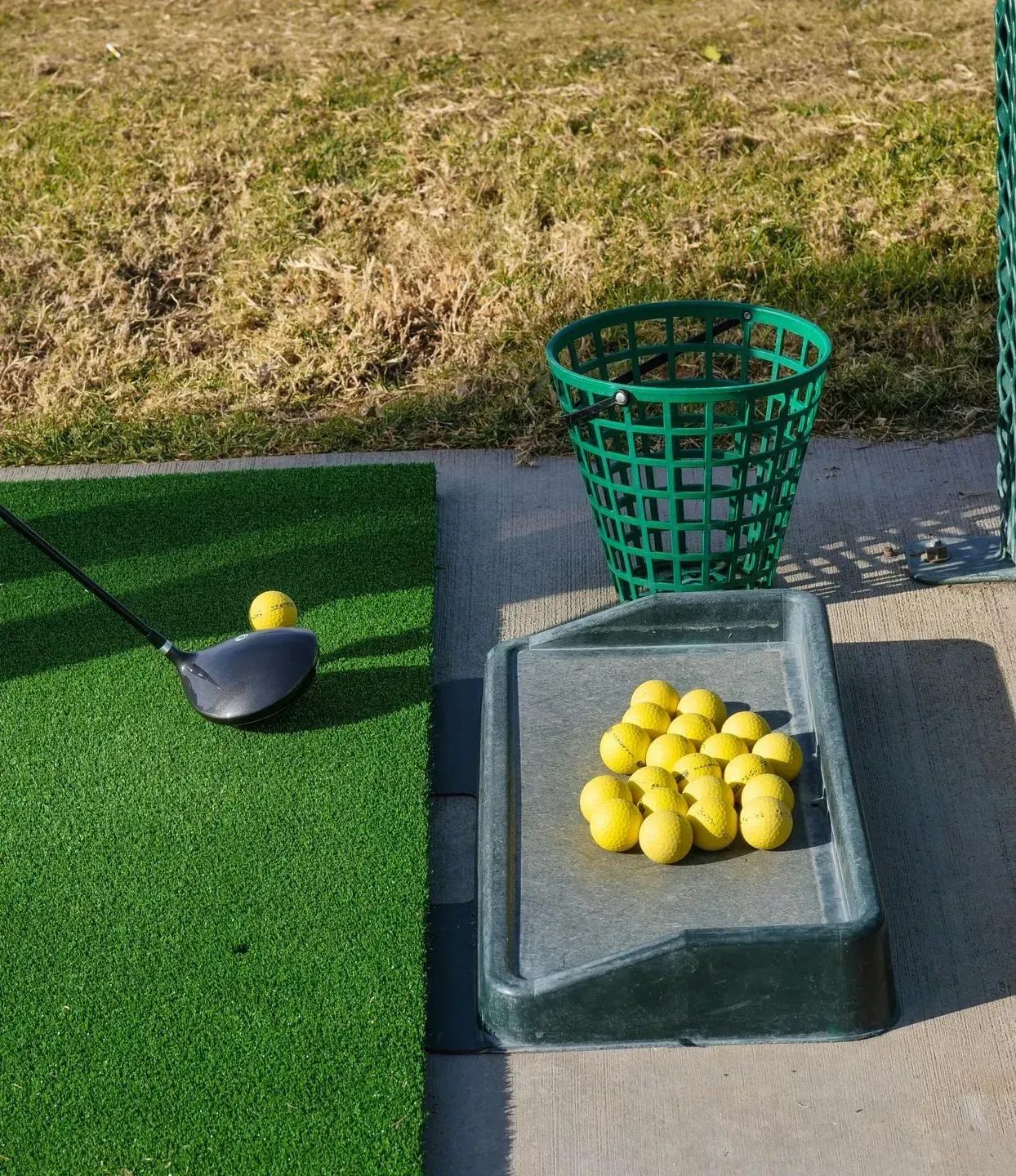 Golf practice mat with a club, golf ball, basket, and a tray of yellow balls on a range.
