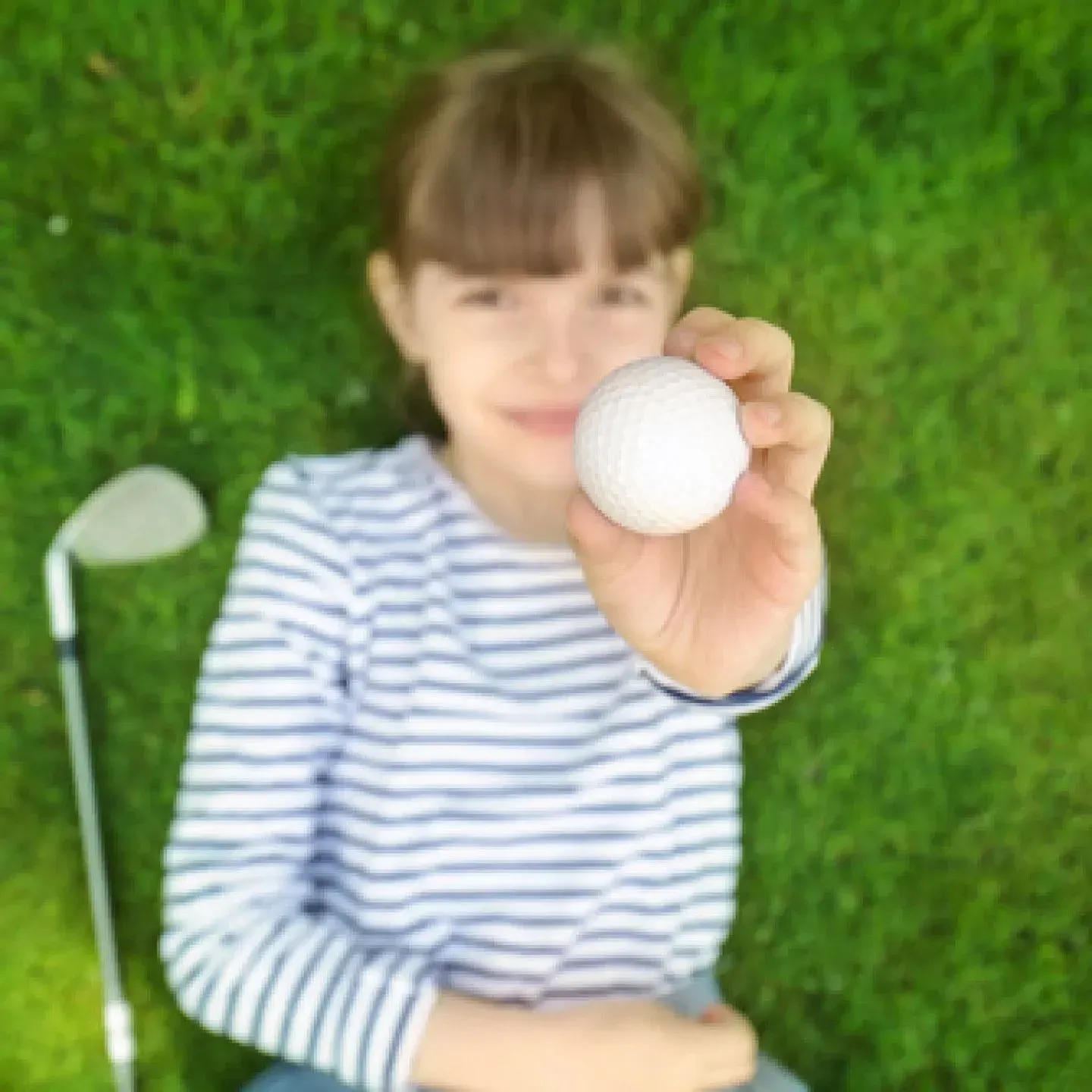 Child holding a golf ball toward the camera on a green putting surface, with a club nearby