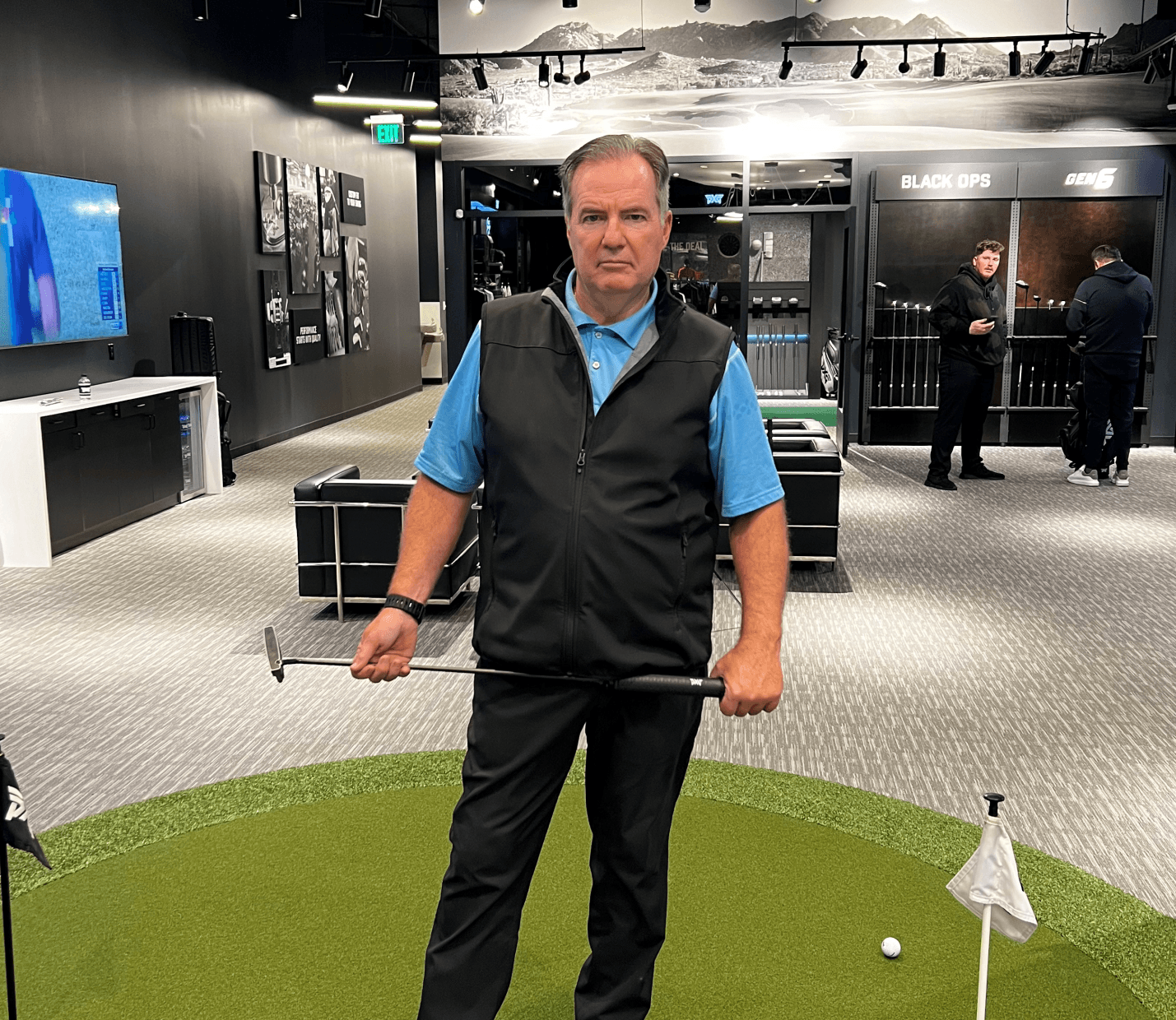 Man holding a golf club in an indoor putting area with green turf and black walls.