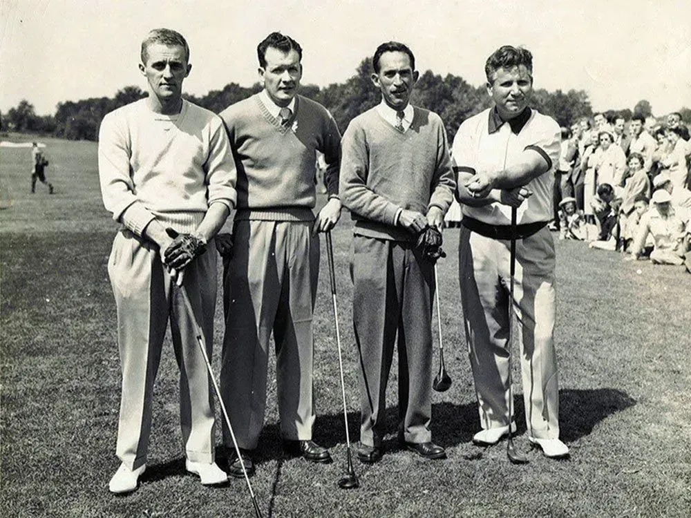 Four golfers posing on a golf course, holding clubs, with a crowd in the background.