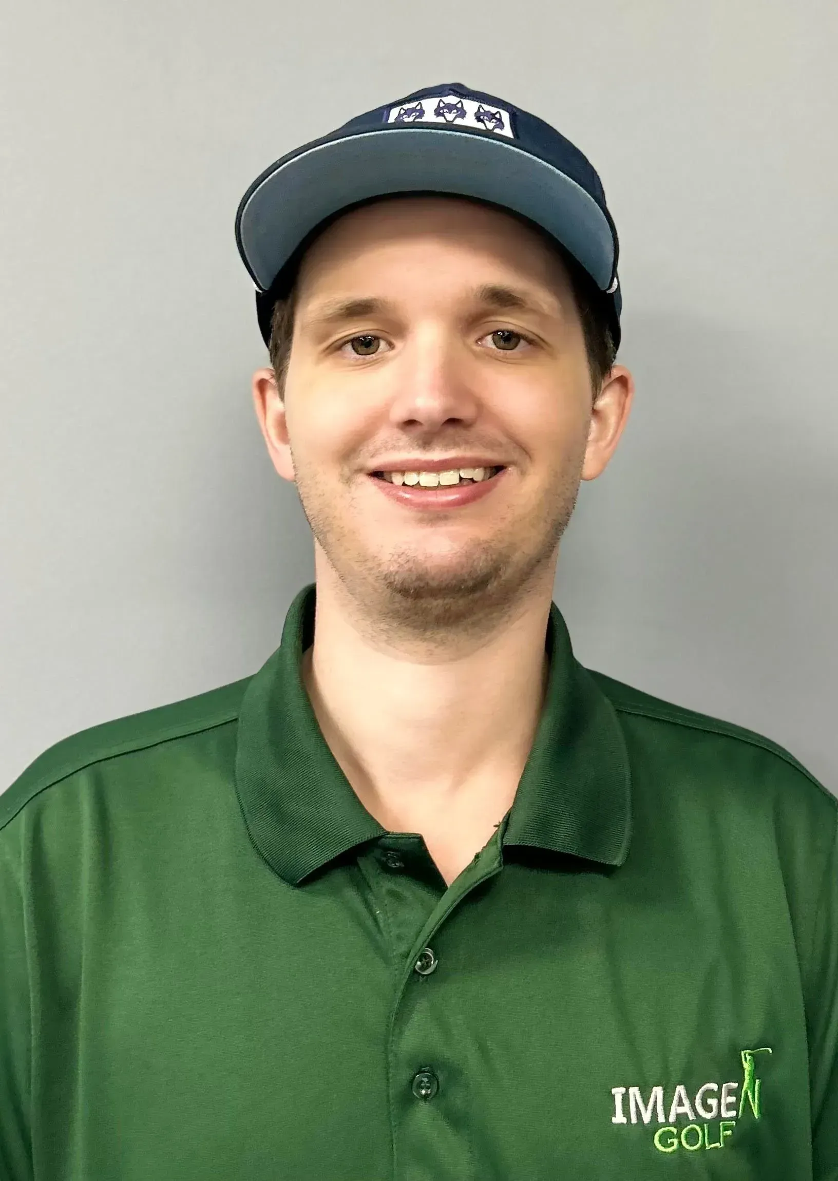 Smiling person in a green polo and blue cap against a gray background.