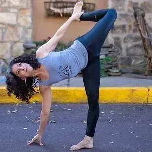 Woman performing a standing yoga pose outdoors on pavement by a stone wall and yellow curb
