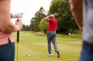 Golfer in red shirt following through on a tee shot on a green course, with spectators watching nearby