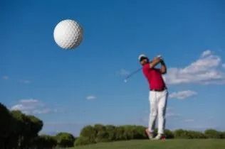 Golfer in red shirt swings a club under a blue sky with a golf ball in the air.