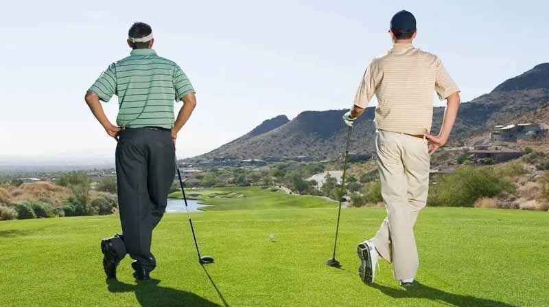 Two golfers seen from behind on a green tee box with clubs, facing a mountainous landscape.