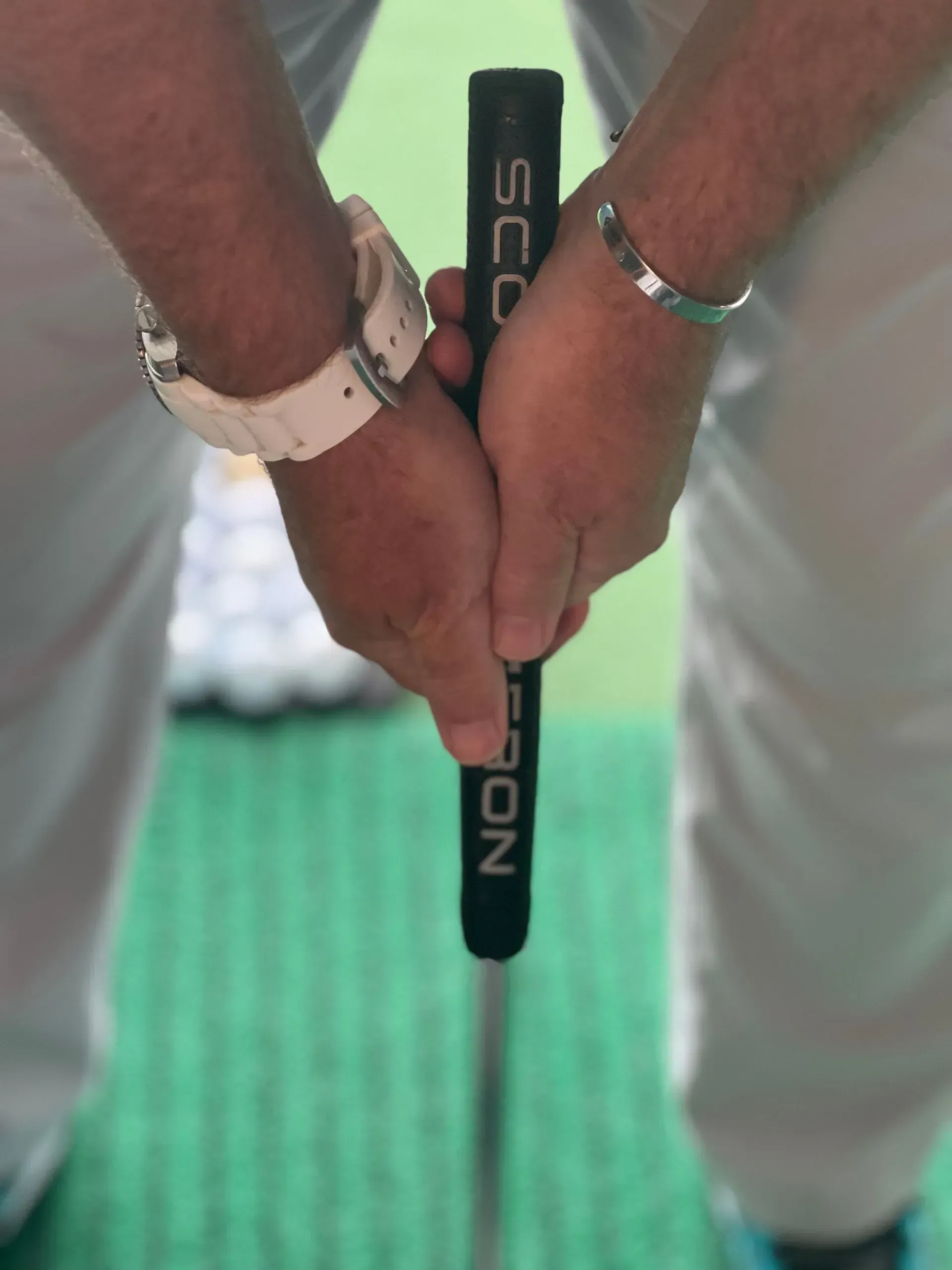 Hands gripping a black golf putter over a green putting surface, with light-colored pants visible.