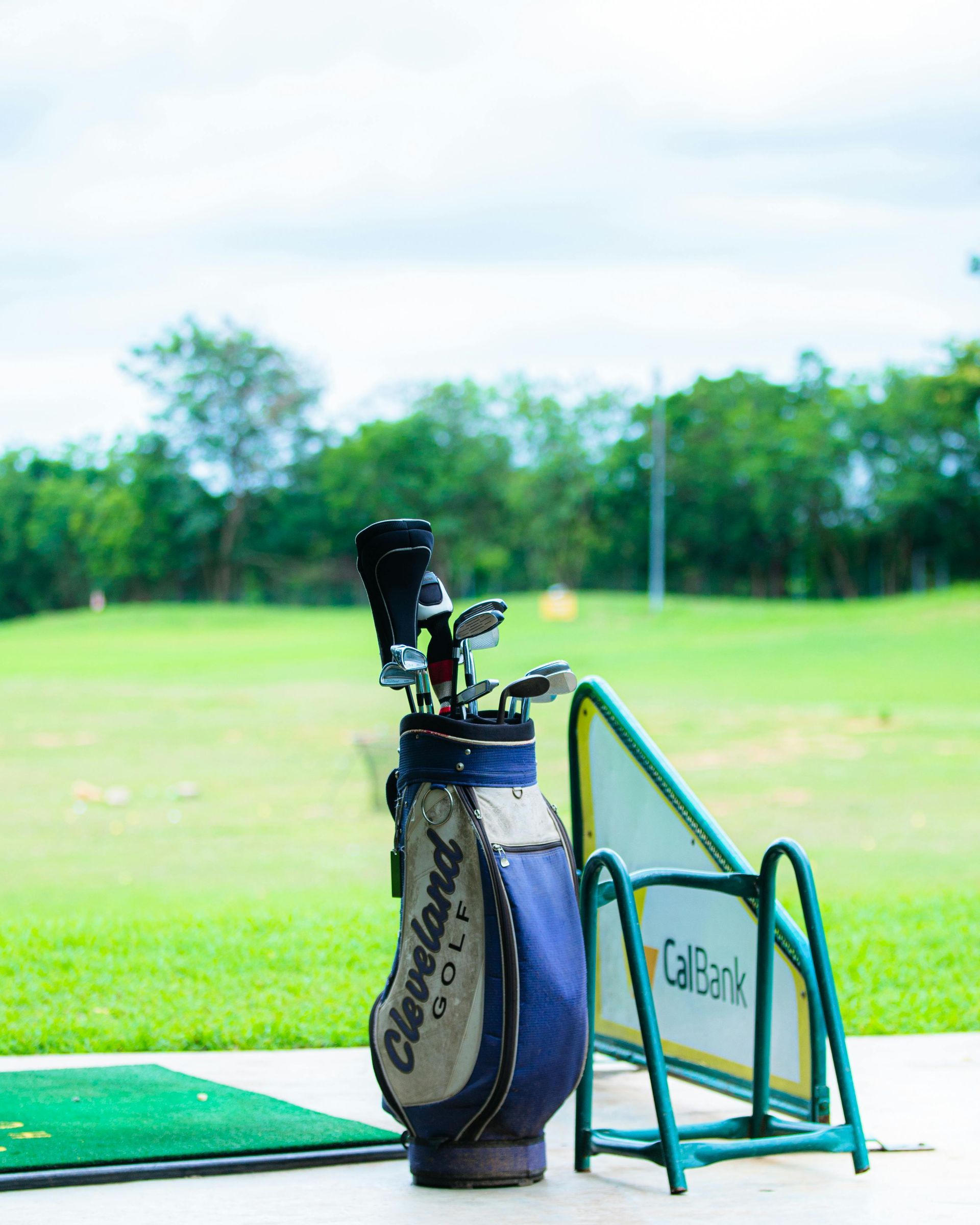 Golf bag with clubs on a cart beside a green driving range under a bright sky