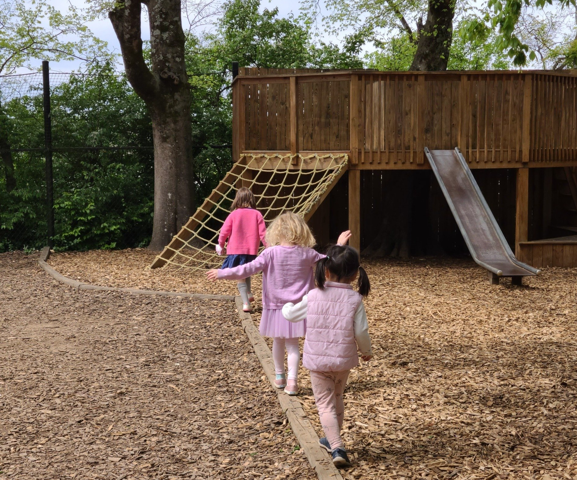 A group of children are walking in line towards a wooden house with a slide