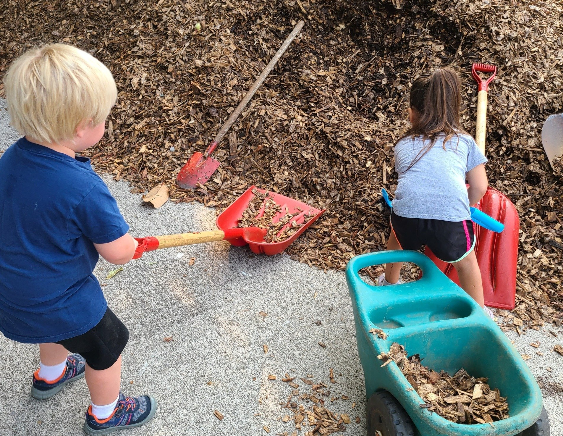 A boy and a girl are playing with shovels and a wheelbarrow full of leaves.
