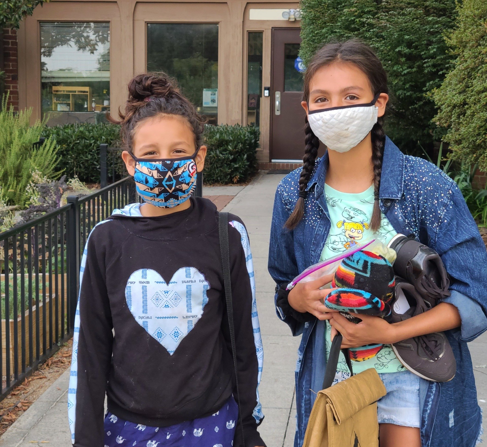 Two girls wearing face masks are standing on a sidewalk