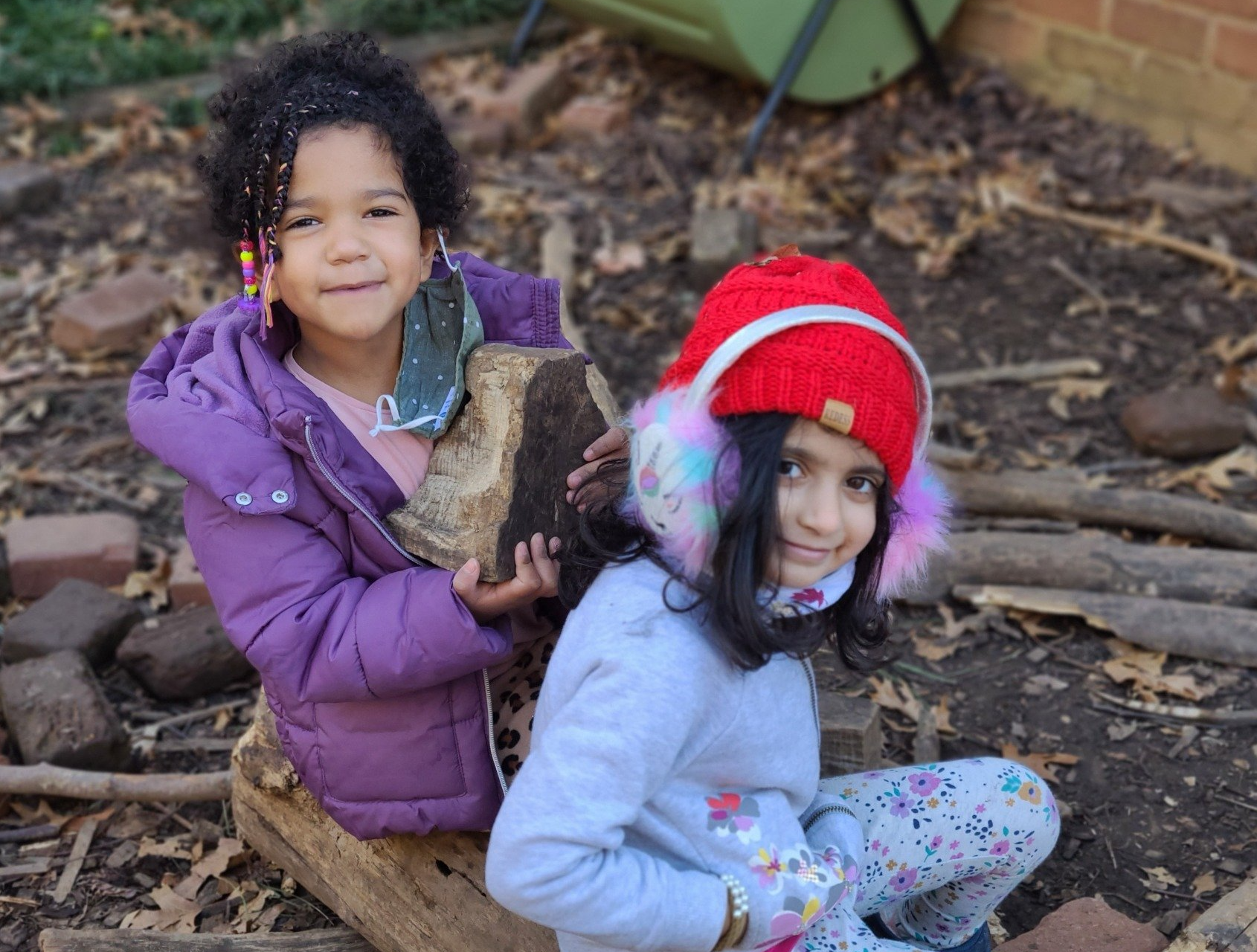Two little girls are sitting on the ground holding logs.