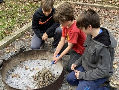 Three young boys are kneeling around a fire pit.