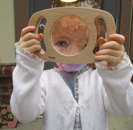 A little girl is holding a wooden montessori material in front of her face.