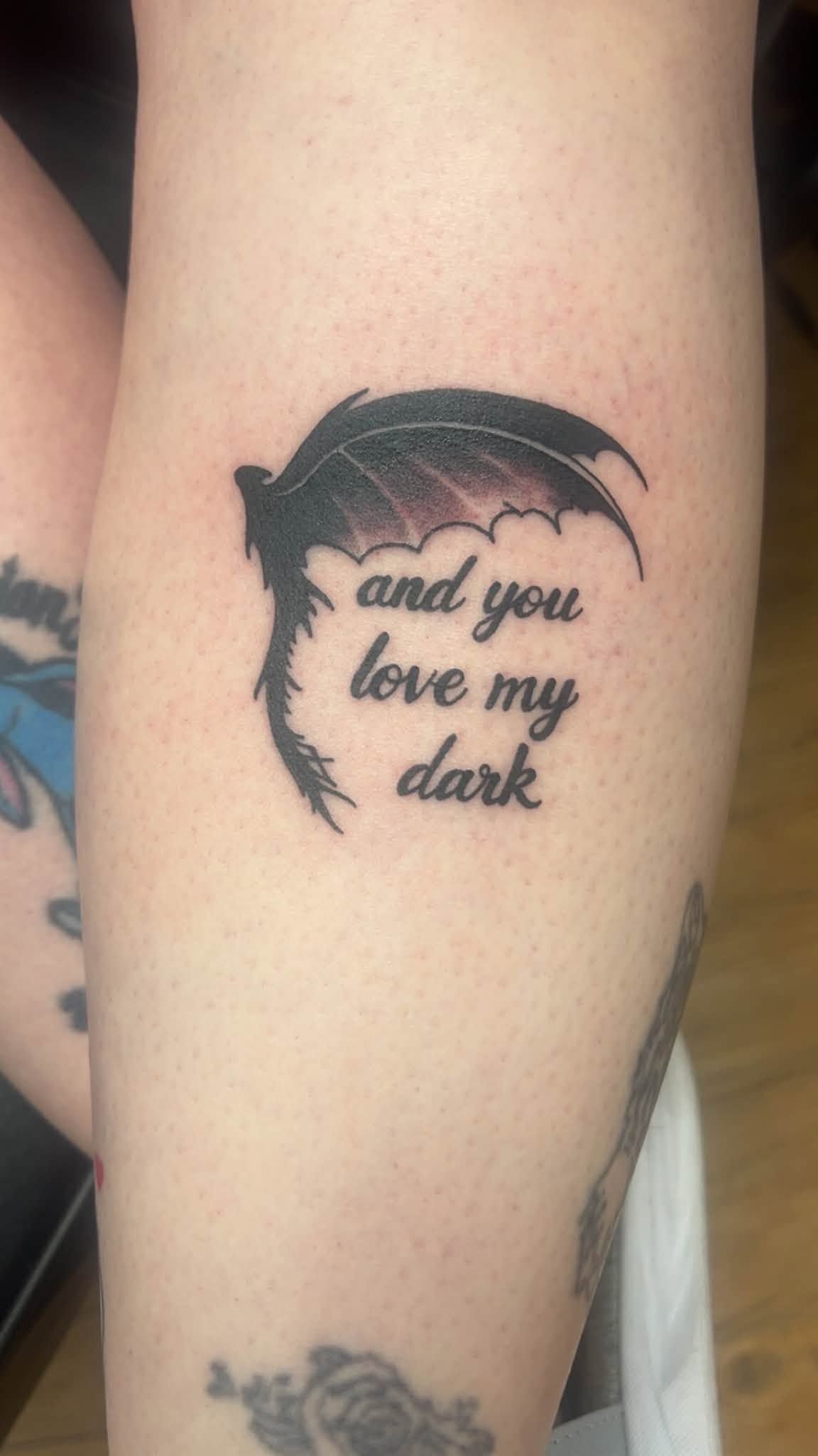 Small black dragon tattoo with the words “and you love my dark” on a forearm