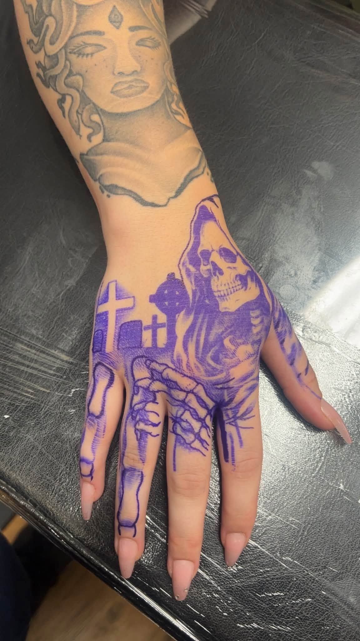 Hand with purple tattoo design of hooded figure and crosses on a black chair
