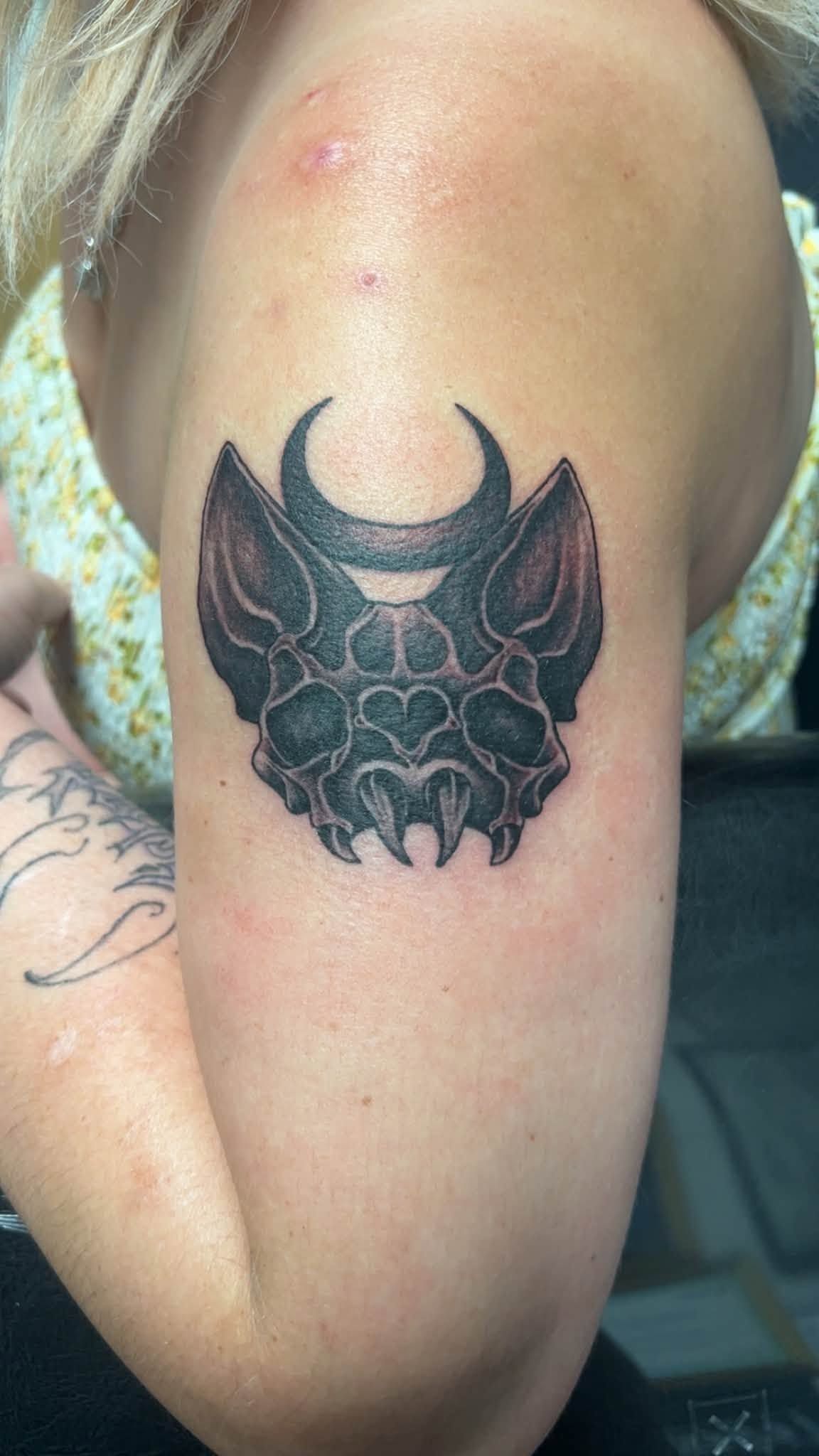 Black bat-like tattoo on a person’s upper arm near the shoulder