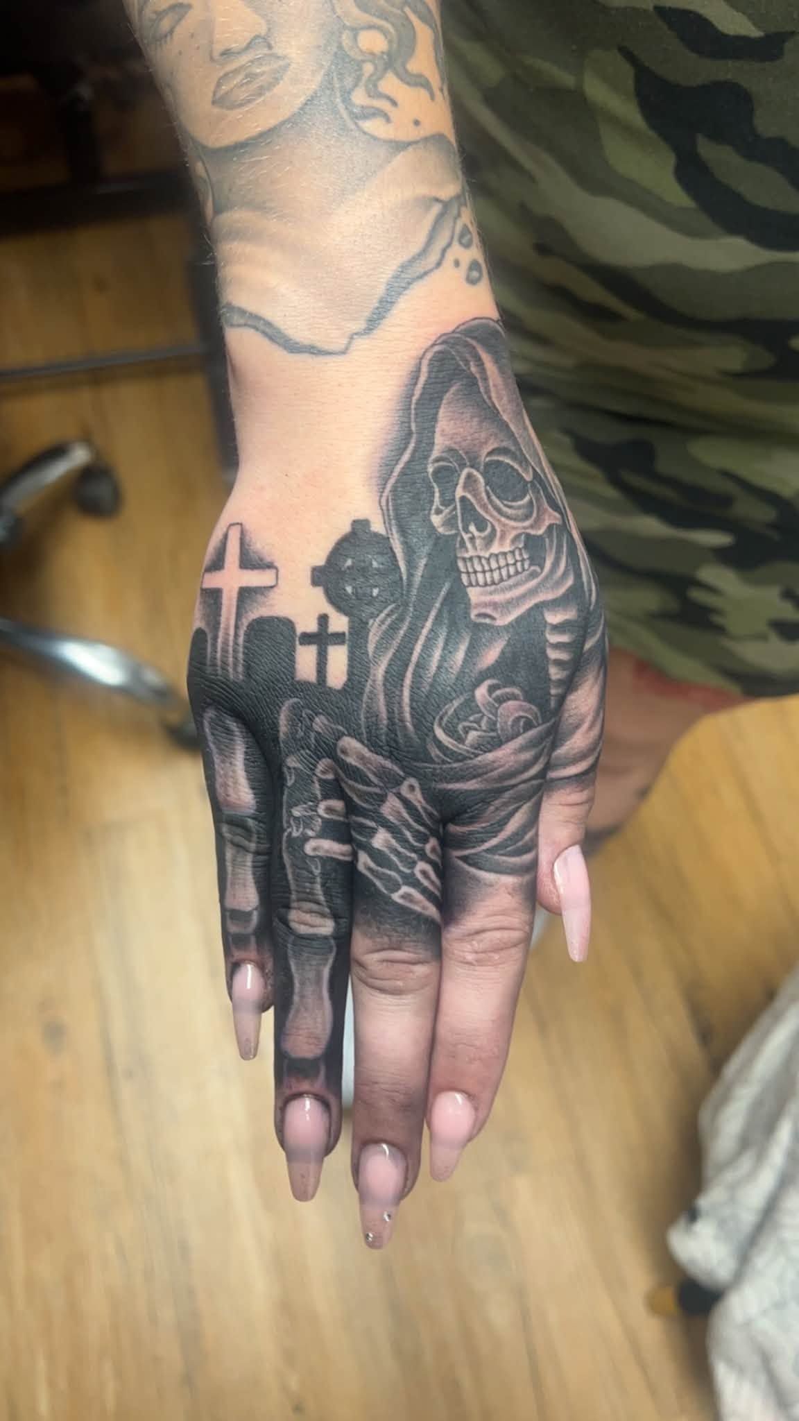 Close-up of two hands with a black tattoo design and long pink nails resting on a wooden floor.