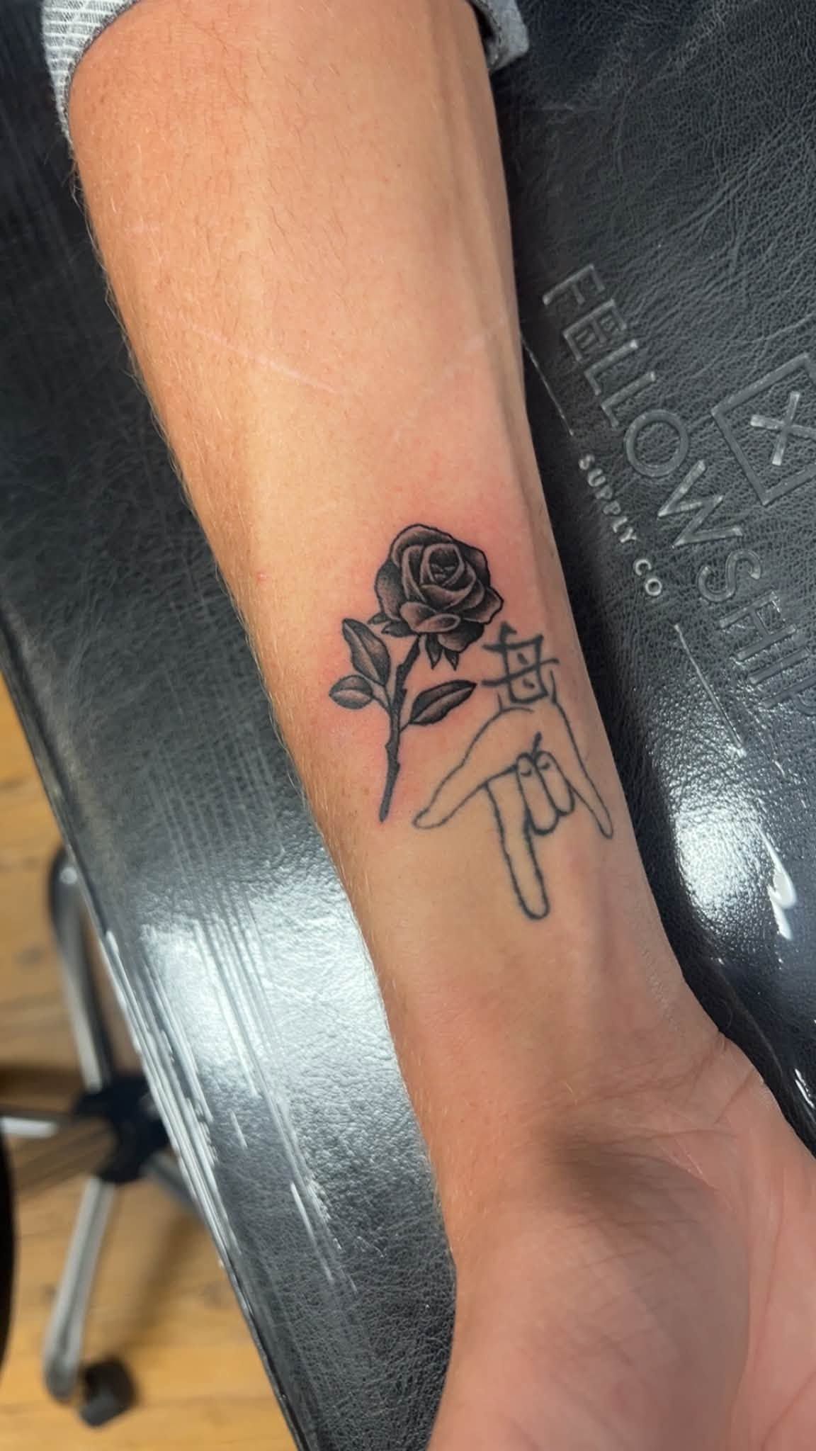Arm with a small black rose and hanging-hand tattoo, resting on a black chair