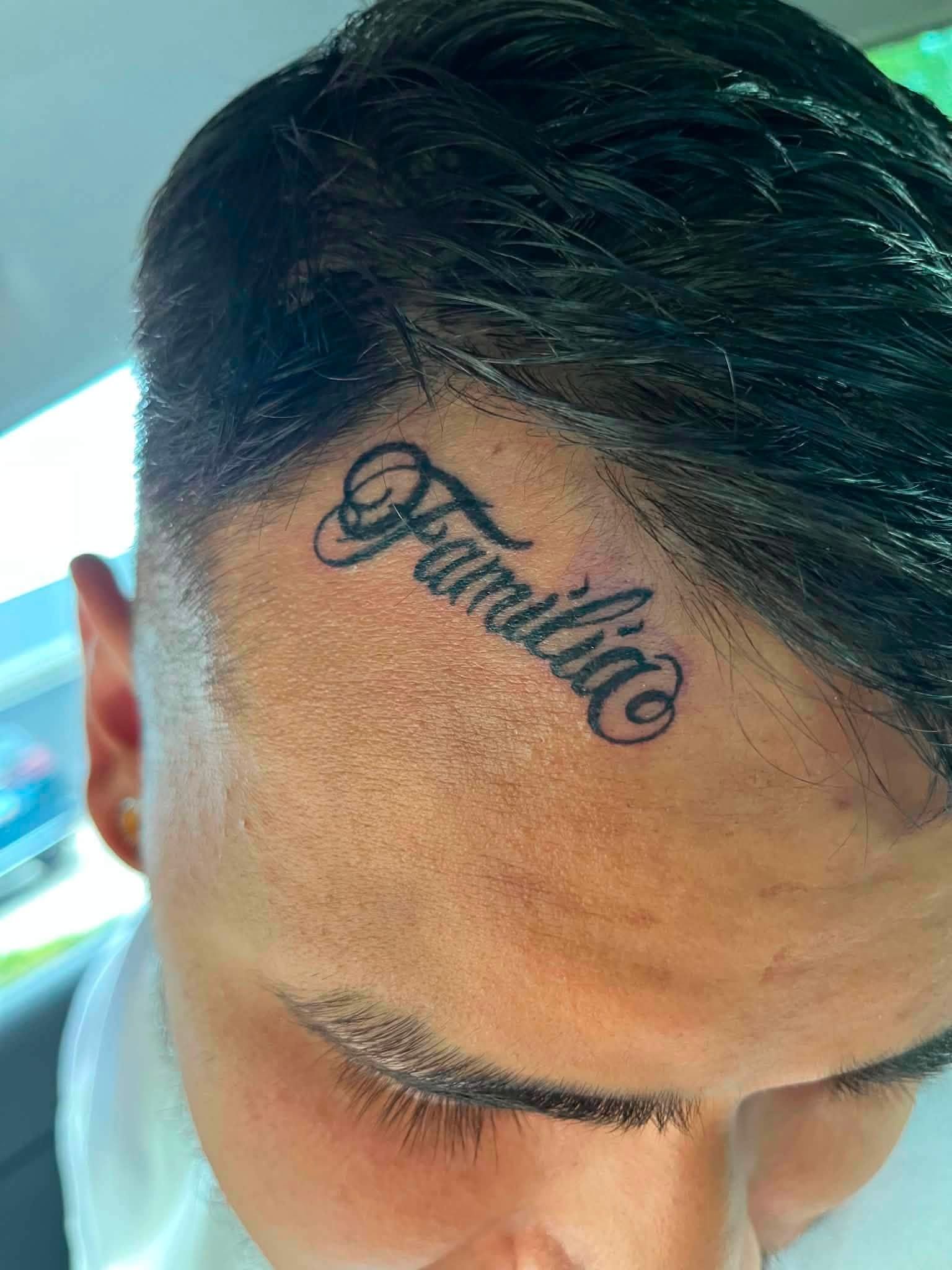 Close-up of a man's forehead with a script tattoo reading “Familia” near the hairline.